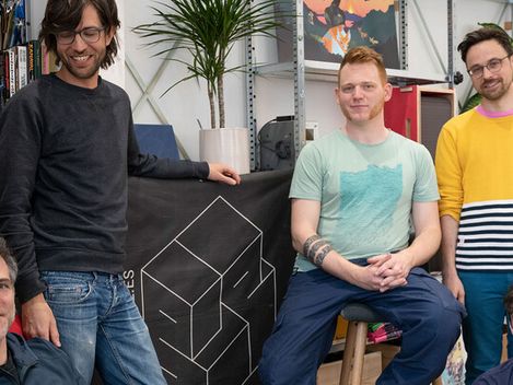 Vier Personen in Freizeitkleidung stehen und sitzen in einem Raum mit Regalen, Pflanzen und Werkzeugen, im Hintergrund ein schwarzes Tuch mit geometrischem Würfelmuster.