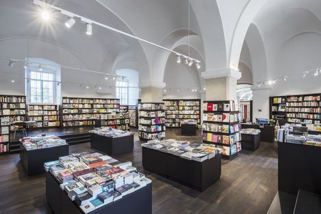 Spacious bookstore with high arched ceilings, filled with neatly organized shelves and tables of books. Bright lighting and a serene atmosphere.