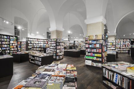 Spacious bookstore with high arched ceilings, filled with neatly organized shelves and tables of books. Bright lighting and a serene atmosphere.