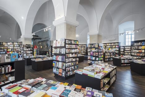 Spacious bookstore with high arched ceilings, filled with neatly organized shelves and tables of books. Bright lighting and a serene atmosphere.