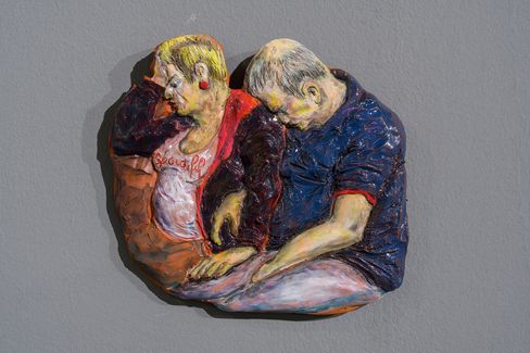 Relief mit zwei sitzenden Personen, die sich an den Köpfen berühren, eine trägt ein weißes Shirt mit rotem Text, die andere ein blaues Hemd.