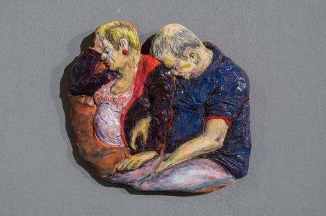 Relief mit zwei sitzenden Personen, die sich an den Köpfen berühren, eine trägt ein weißes Shirt mit rotem Text, die andere ein blaues Hemd.