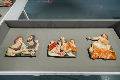 Drei farbige Reliefs mit menschlichen Figuren in verschiedenen Posen auf einer grauen Ausstellungsfläche.