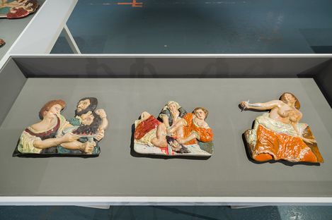 Drei farbige Reliefs mit menschlichen Figuren in verschiedenen Posen auf einer grauen Ausstellungsfläche.