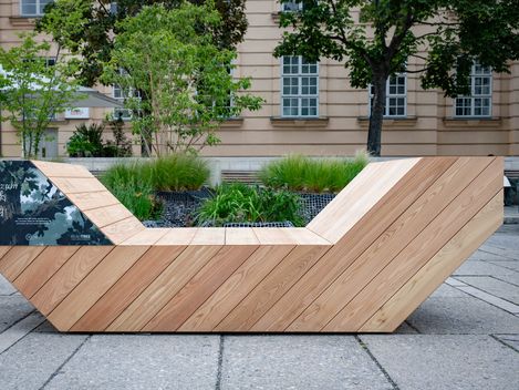 Wooden Enzi seating element at MuseumsQuartier Vienna, set among planted beds in front of a historic building façade.