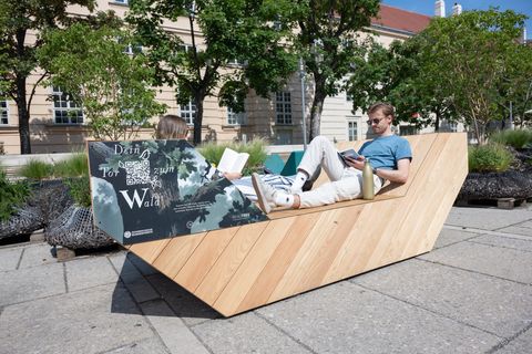 Two people relax and read on a wooden Enzi seating element at MuseumsQuartier, surrounded by greenery and historic buildings.