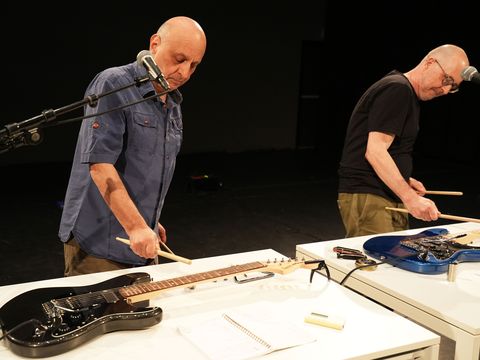 Die Künstler spielen mit Drumsticks auf E-Gitarren, die auf zwei Tischen vor ihnen liegen.