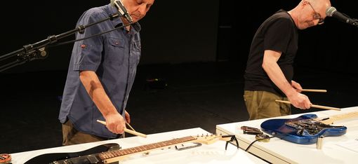 Die Künstler spielen mit Drumsticks auf E-Gitarren, die auf zwei Tischen vor ihnen liegen.