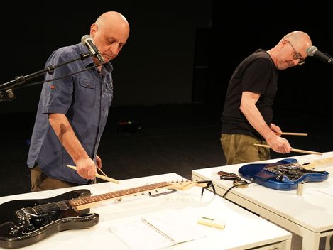 Die Künstler spielen mit Drumsticks auf E-Gitarren, die auf zwei Tischen vor ihnen liegen.
