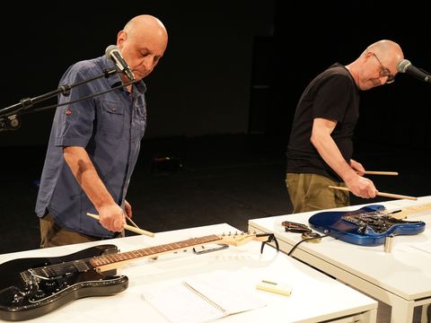 Die Künstler spielen mit Drumsticks auf E-Gitarren, die auf zwei Tischen vor ihnen liegen.