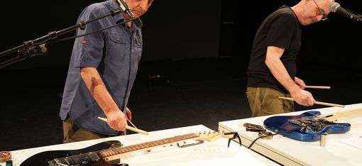 Die Künstler spielen mit Drumsticks auf E-Gitarren, die auf zwei Tischen vor ihnen liegen.