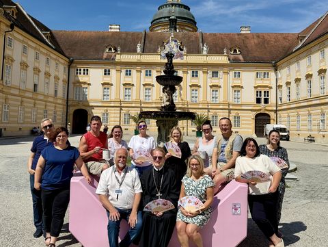 Group photograph in front of the Baroque Melk Abbey: Several people are seated and standing around a pink Enzi beside a fountain in the courtyard. The historic monastery complex with its distinctive dome rises in the background beneath a blue sky.
