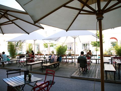 Outdoor cafe patio with wooden tables and red chairs under large white umbrellas. People are seated, engaged in conversation, surrounded by potted plants.