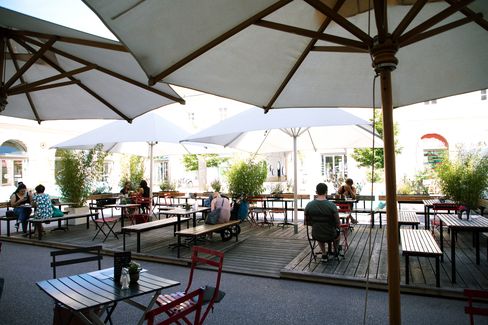 Outdoor cafe patio with wooden tables and red chairs under large white umbrellas. People are seated, engaged in conversation, surrounded by potted plants.
