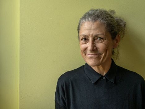 Frau mit zusammengebundenem Haar und schwarzem Pullover steht vor einer hellgrünen Wand und lächelt leicht
