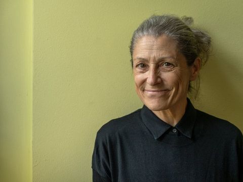 Woman with tied-back hair wearing a black sweater standing against a light green wall with a slight smile