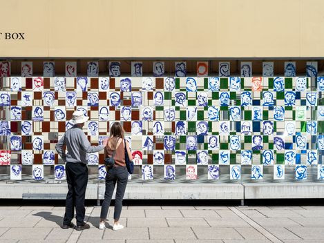 Two people stand in front of a wall installation made of square tiles with abstract, graphic faces and patterns.