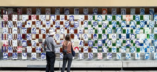 Two people stand in front of a wall installation made of square tiles with abstract, graphic faces and patterns.