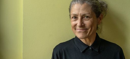 Woman with tied-back hair wearing a black sweater standing against a light green wall with a slight smile