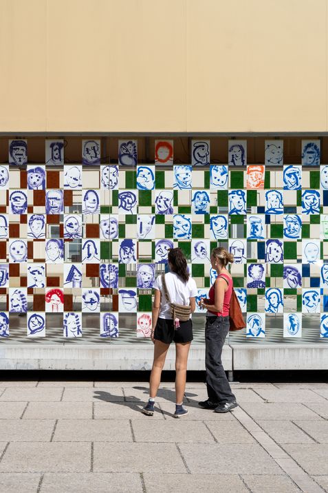 Two women stand in front of a wall covered with a checkerboard pattern of square images showing various faces in blue, red, and black and white