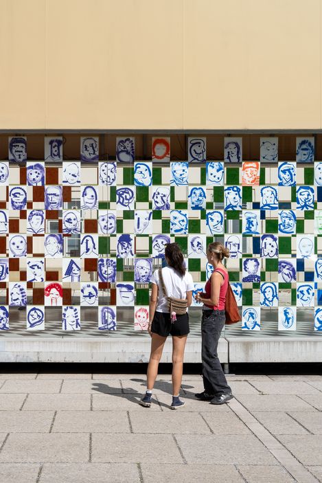 Two women stand in front of a wall covered with a checkerboard pattern of square images showing various faces in blue, red, and black and white