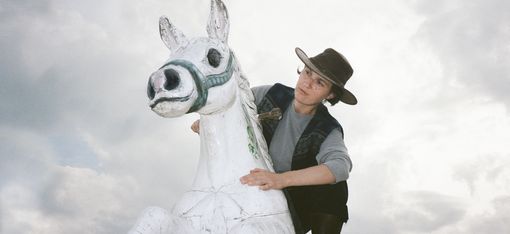 Kind mit Cowboyhut und Weste sitzt auf einer großen weißen Pferdeskulptur vor bewölktem Himmel