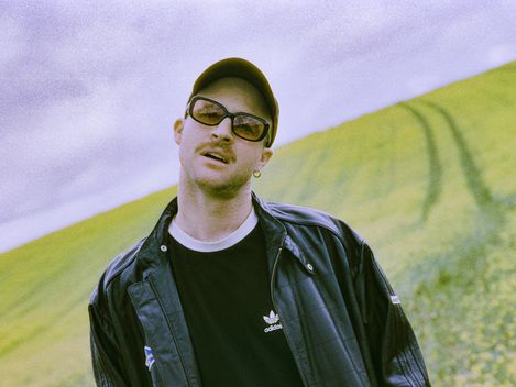 Young man wearing a black Adidas jacket and black T-shirt standing in front of a hilly landscape