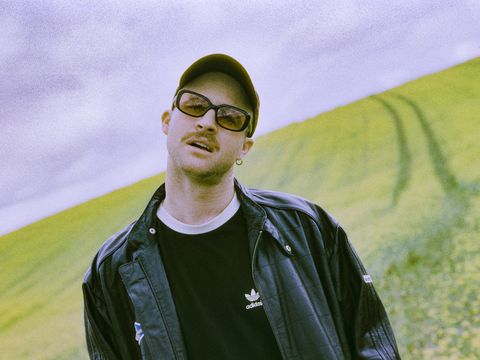 Young man wearing a black Adidas jacket and black T-shirt standing in front of a hilly landscape