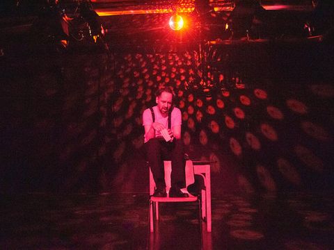The actor is sitting on the back of an armchair. He is holding a cone of popcorn. The stage is lit in red.