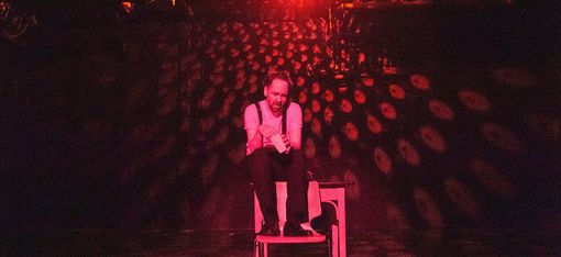 The actor is sitting on the back of an armchair. He is holding a cone of popcorn. The stage is lit in red.