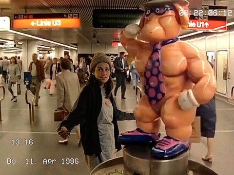 Frau mit Mütze und dunkler Jacke steht in einer U-Bahn-Station neben einer übergroßen, muskulösen Cartoonfigur mit Krawatte und Handschuhen