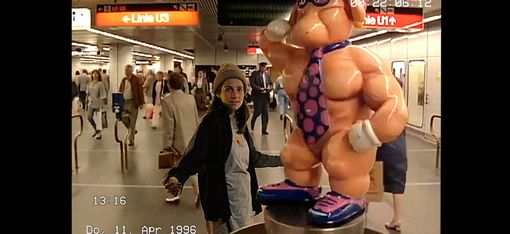 Frau mit Mütze und dunkler Jacke steht in einer U-Bahn-Station neben einer übergroßen, muskulösen Cartoonfigur mit Krawatte und Handschuhen
