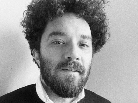 Person with curly hair, beard, and sweater over shirt against plain background.