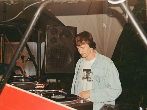Person wearing headphones stands at DJ booth with turntables and speaker in the background.