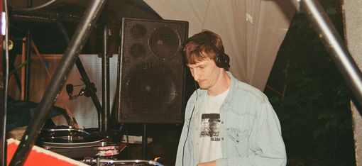 Person wearing headphones stands at DJ booth with turntables and speaker in the background.
