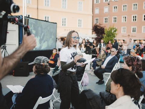 Frau mit Brille und Mikrofon spricht vor sitzendem Publikum im Freien vor einem großen Bildschirm