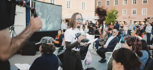 Frau mit Brille und Mikrofon spricht vor sitzendem Publikum im Freien vor einem großen Bildschirm