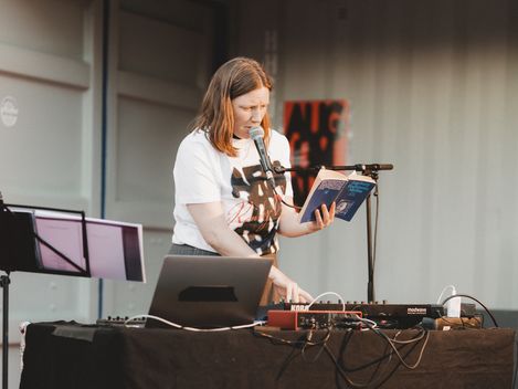 Frau steht hinter einem Tisch mit elektronischer Musik-Ausrüstung, hält ein Buch und spricht in ein Mikrofon
