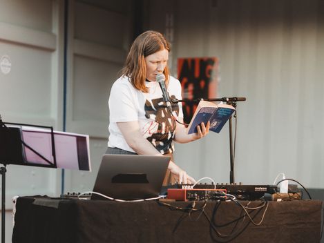 Frau steht hinter einem Tisch mit elektronischer Musik-Ausrüstung, hält ein Buch und spricht in ein Mikrofon