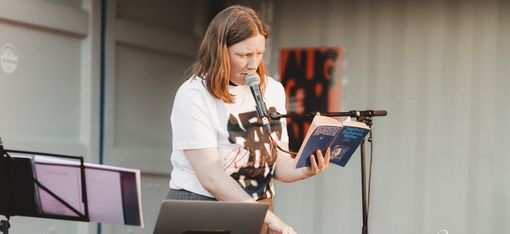 Frau steht hinter einem Tisch mit elektronischer Musik-Ausrüstung, hält ein Buch und spricht in ein Mikrofon