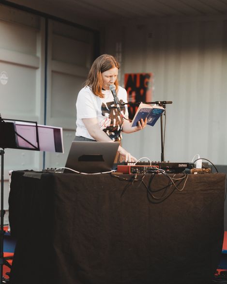 Frau steht hinter einem Tisch mit elektronischer Musik-Ausrüstung, hält ein Buch und spricht in ein Mikrofon