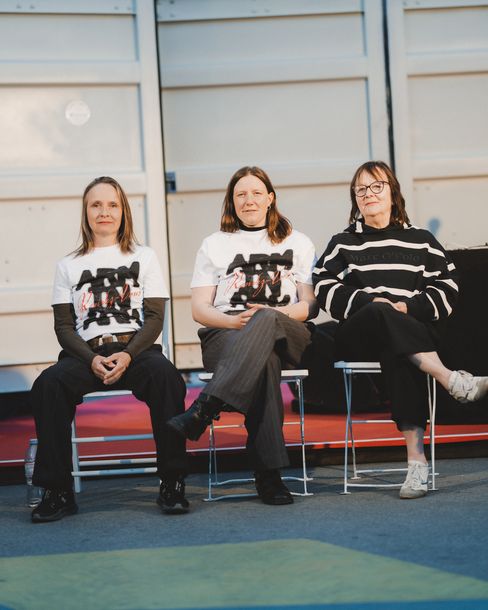 Drei Frauen sitzen nebeneinander auf Stühlen vor einer hellen Wand, zwei tragen weiße T-Shirts mit schwarzem und rotem Aufdruck, die dritte trägt einen schwarz-weißen Pullover mit Streifen.
