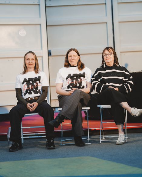 Drei Frauen sitzen nebeneinander auf Stühlen vor einer hellen Wand, zwei tragen weiße T-Shirts mit schwarzem und rotem Aufdruck, die dritte trägt einen schwarz-weißen Pullover mit Streifen.