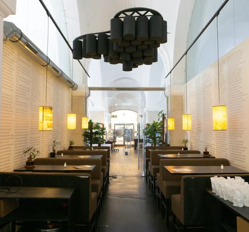 A long, bright hallway with dark wooden tables and benches on either side. Warm lights hang from the walls.