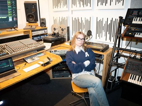 Person with crossed arms sitting on a swivel chair in a recording studio surrounded by mixing consoles, speakers, synthesizers, and acoustic panels.