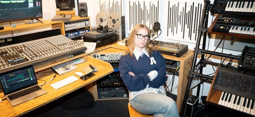 Person with crossed arms sitting on a swivel chair in a recording studio surrounded by mixing consoles, speakers, synthesizers, and acoustic panels.