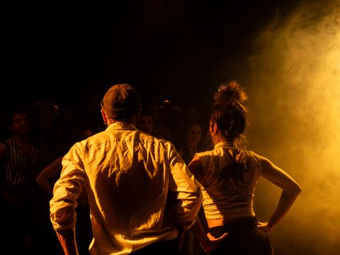 Two people seen from behind with hands on hips in front of bright light in dark setting.