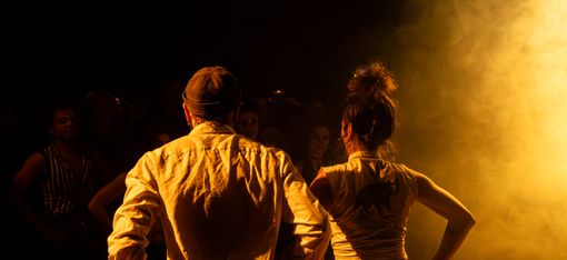 Two people seen from behind with hands on hips in front of bright light in dark setting.