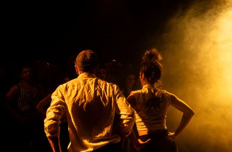 Zwei Personen von hinten mit Händen in der Hüfte vor einem hellen Licht in dunkler Umgebung.