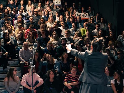 Das Foto zeigt Menschen, die im Publikum sitzen. Sie schauen auf eine Frau, die auf der Bühne steht. Neben ihr steht ein Mikrofon.